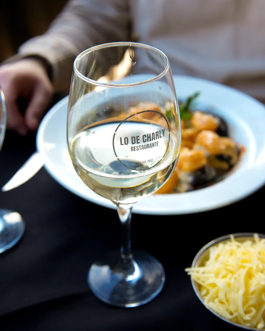 Copa de vino blanco con el logo de Lo de Charly, al lado de un plato de pasta con mariscos